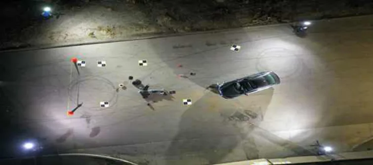 Overhead night view of the staged accident site lit by scene lights, showing the black car, crash dummy, cones, measuring tape, and checkerboard ground control point markers